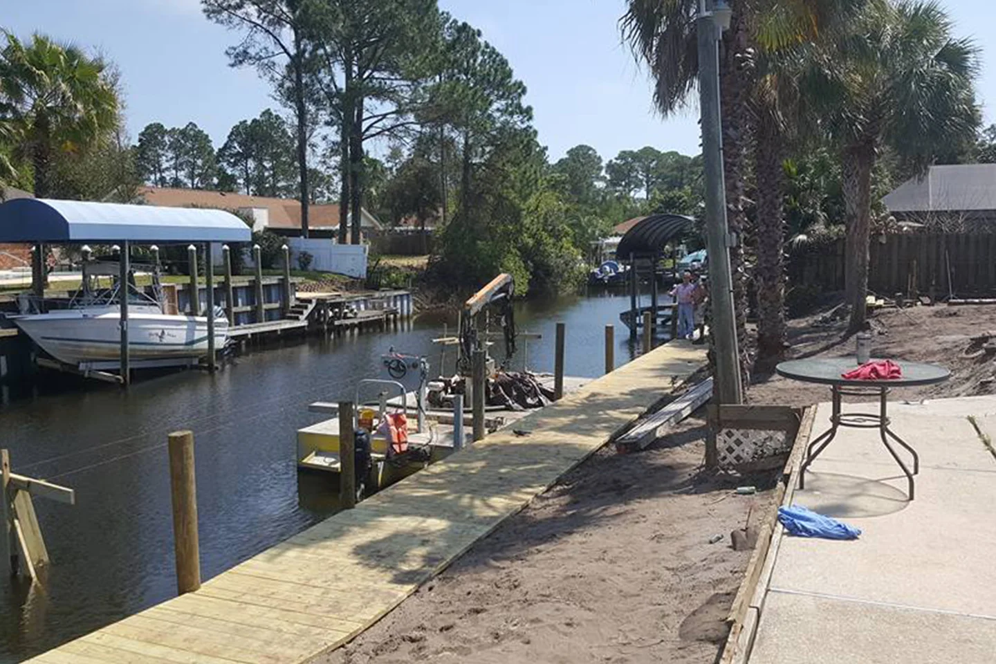 A wooden dock extends along a narrow canal, where boats are moored. A few palm trees line the path, and some construction equipment is visible. A patio area with a table is on the right.