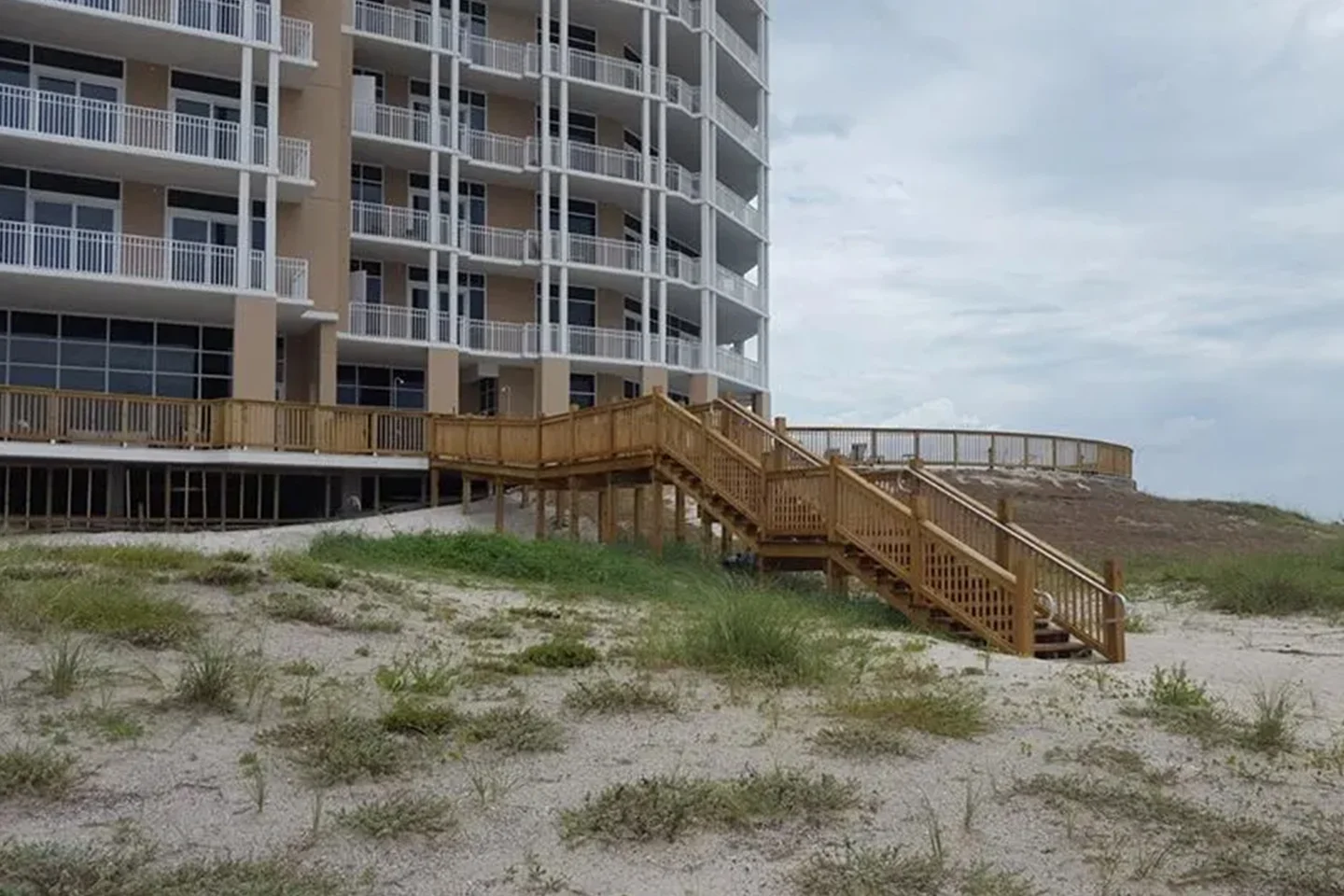 A beachfront condomium with balconies is connected to the sandy beach area by a wooden staircase and boardwalk. The surrounding area has sparse vegetation. The sky is overcast.