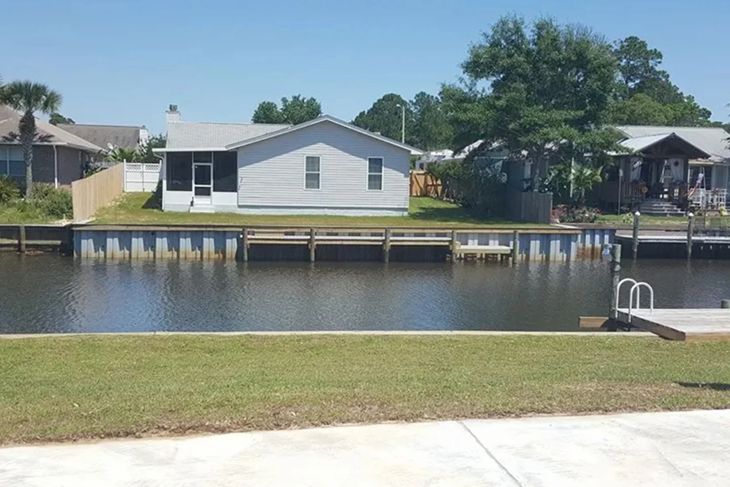 A small gray house with a fenced backyard sits by a canal, with a dock and ladder leading to the water and a large tree to the right.