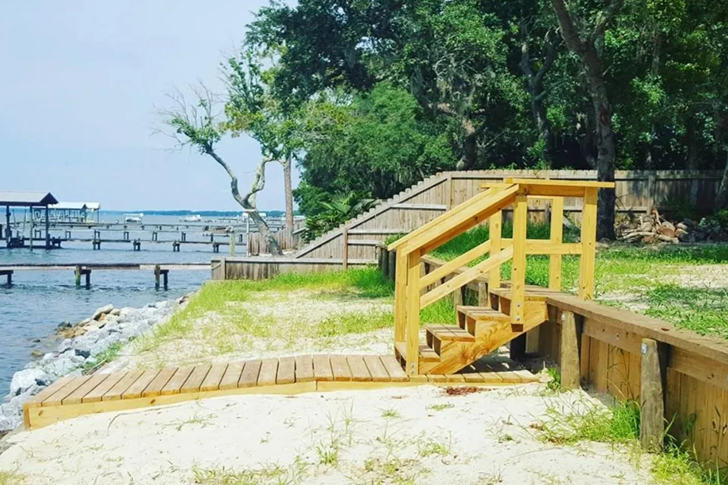 A small wooden staircase and boardwalk lead to a sandy beach with grass and trees nearby. Piers extend into the water in the background.