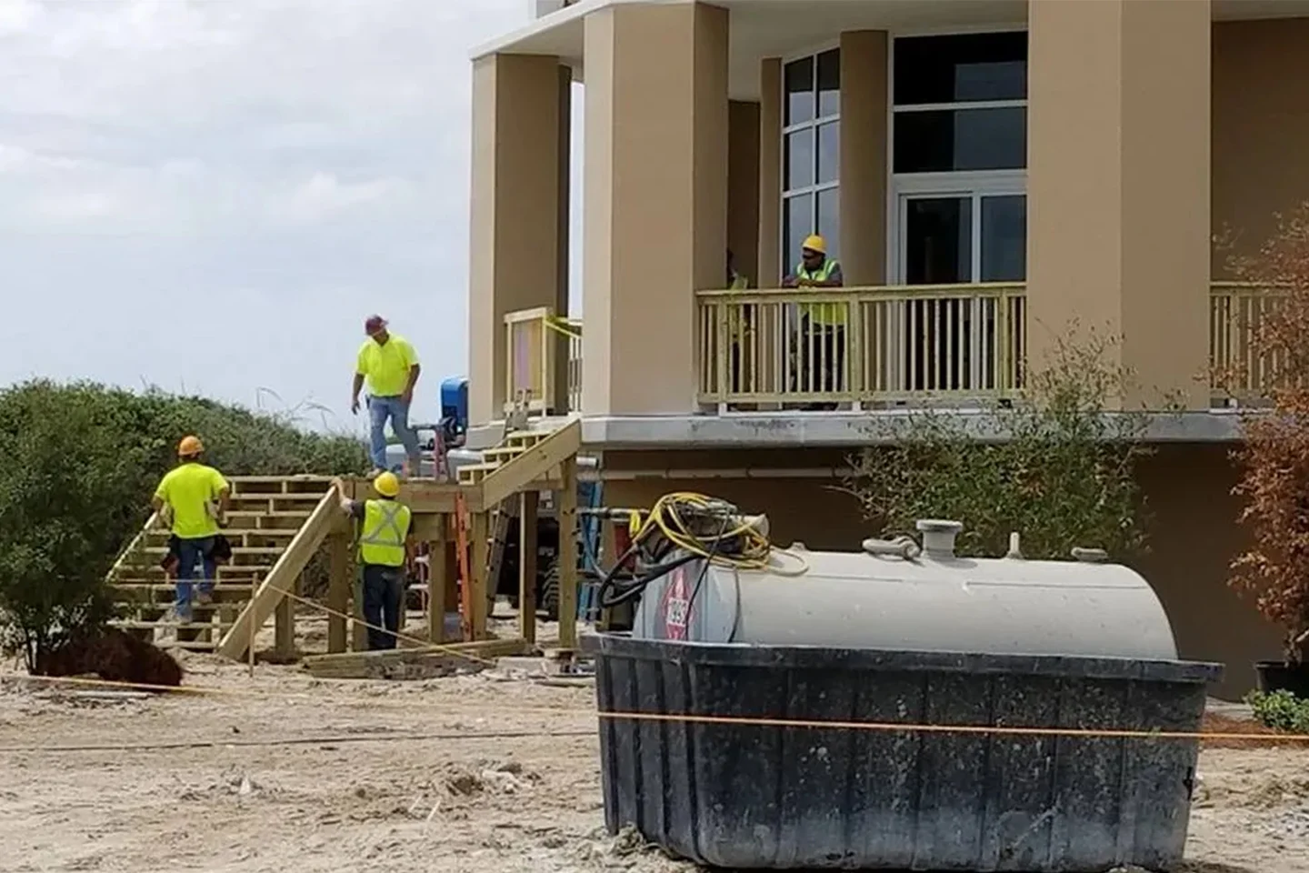 Workers in high-visibility vests and helmets construct a staircase and porch on a modern two-story building, with construction materials and equipment visible in the foreground.