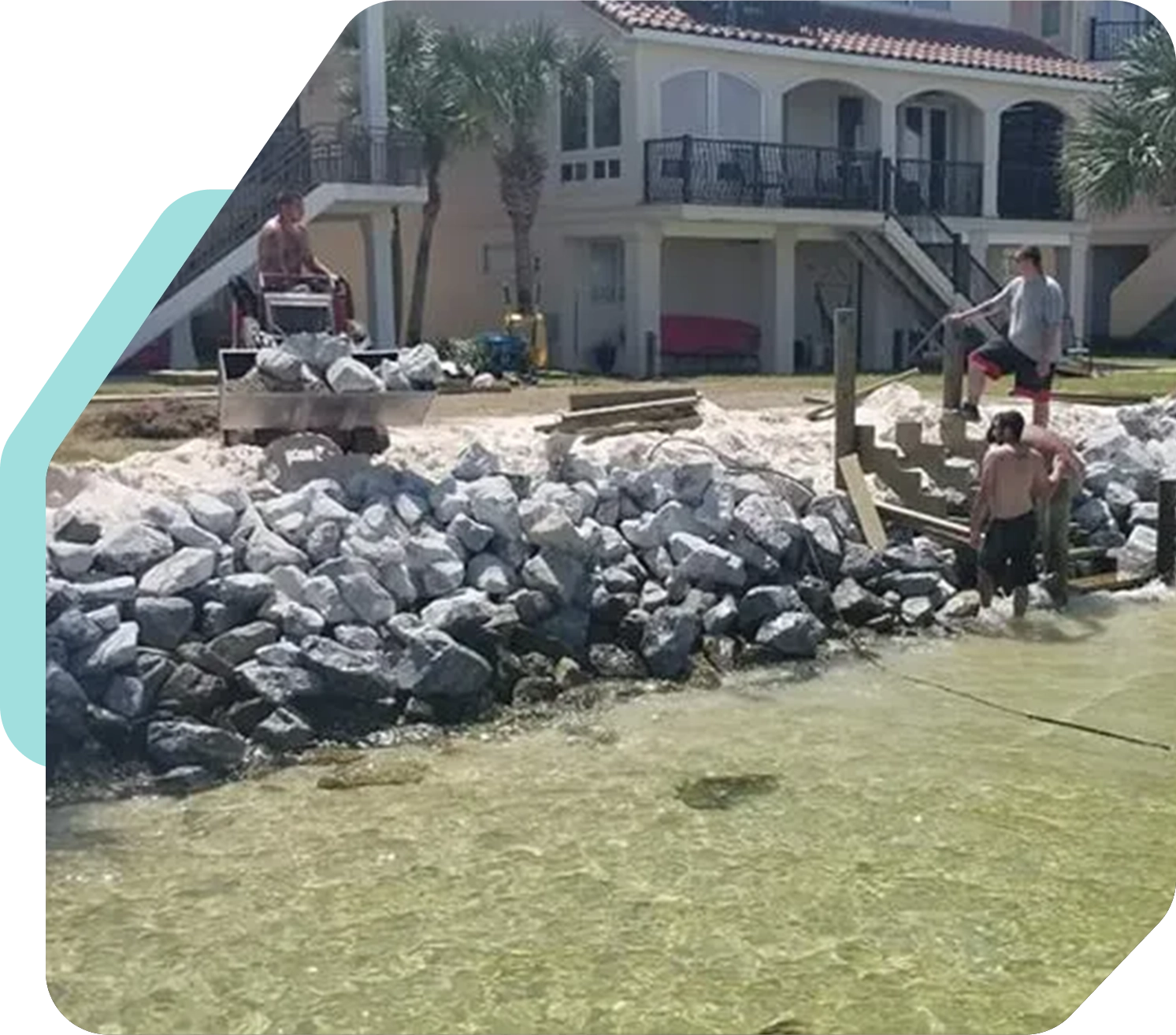 Three workers are constructing a seawall made of large rocks in front of a waterfront property. One worker operates a machine while the other two position rocks.